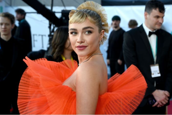 Florence Pugh looks at the camera while wearing an off-the-shoulder orange dress with big, puffy ruffles. 