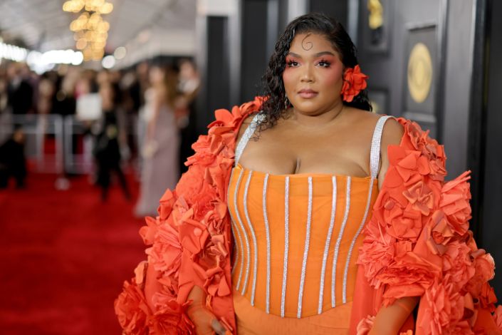 Lizzo arrives to the Grammys wearing an orange dress with a matching orange shoulder wrap and flower in her hair. 