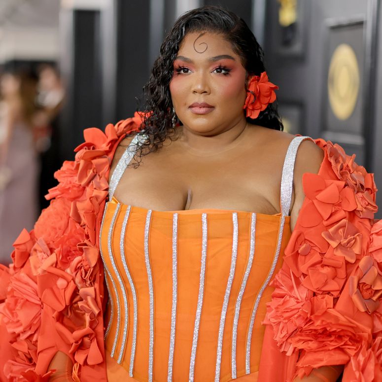 Lizzo arrives to the Grammys wearing an orange dress with a matching orange shoulder wrap and flower in her hair.
