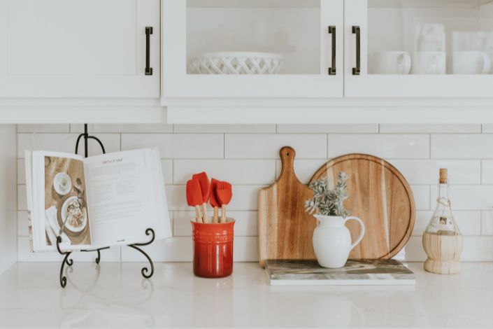 A photo of a white kitchen counter that features an open cookbook and common kitchen tools. 