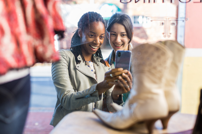 Two smiling friends snap a picture of shoes while window-shopping 