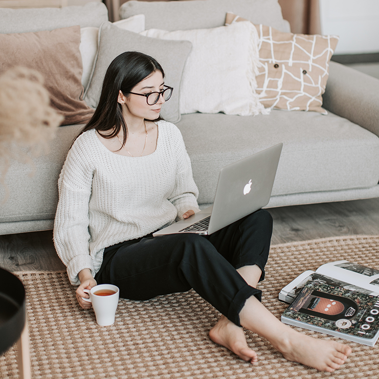 a young white woman with glasses sitting on the floor of her living room looking at her laptop